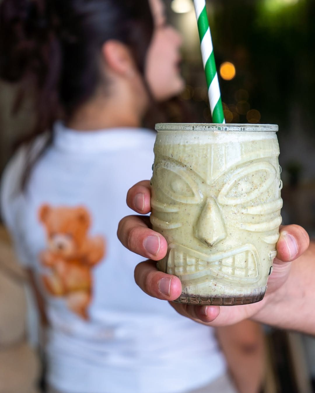 Mão a segurar um copo tiki com uma bebida verde-clara e uma palhinha às riscas