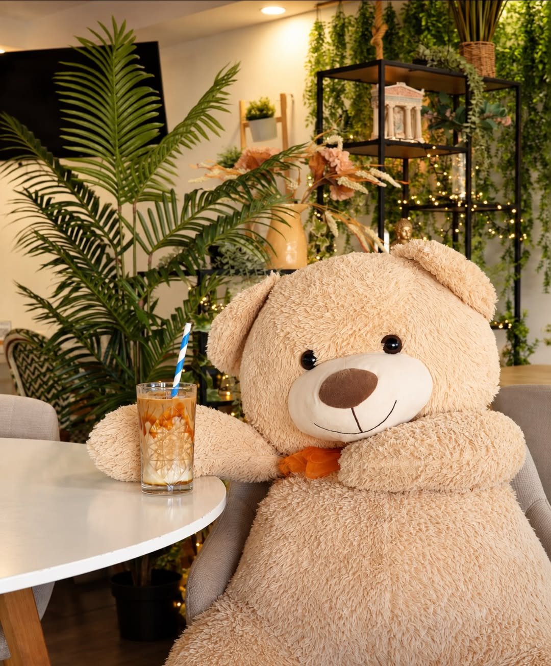 Urso de peluche gigante sentado a uma mesa branca, com um copo com uma bebida e palhinha, plantas e luzes ao fundo