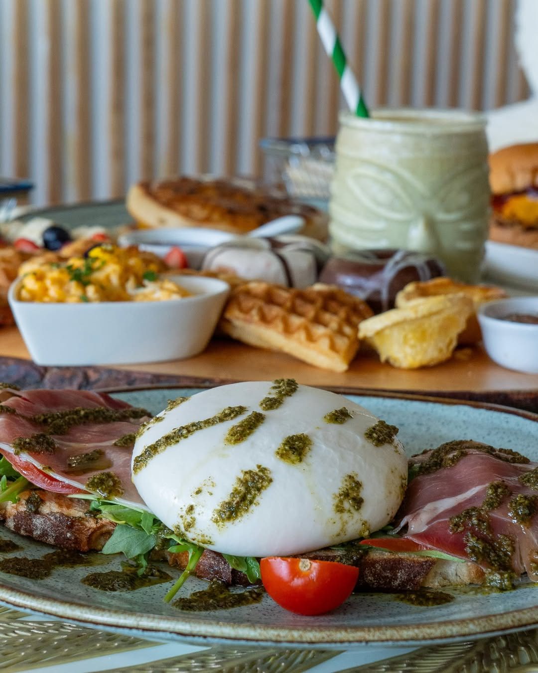 Burrata com pesto e presunto num prato azul, com waffles, ovos mexidos e um copo de batido ao fundo
